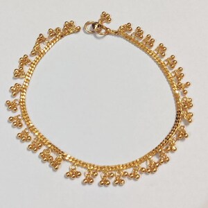 May include: A gold-colored anklet with a delicate chain design. The anklet features small, dangling floral and ball-shaped charms. The chain is approximately 9 inches in length, with a clasp closure.