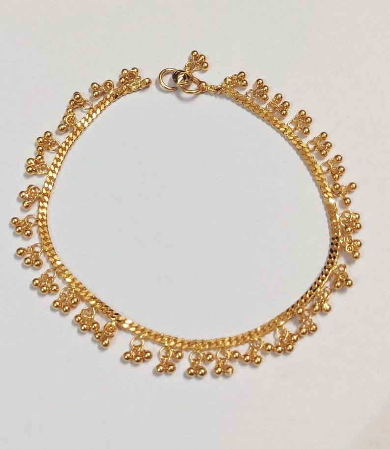 May include: A gold-colored anklet with a delicate chain design. The anklet features small, dangling floral and ball-shaped charms. The chain is approximately 9 inches in length, with a clasp closure.