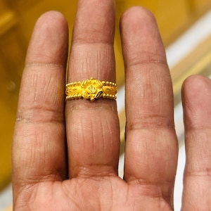 Op de afbeelding: Een gouden ring met een bloemmotief wordt op een vinger getoond. De ring heeft een centrale bloem met gedetailleerde bloemblaadjes en kralenbanden. Het ingewikkelde ontwerp en de heldere goudkleur worden uitgelicht in deze close-up.