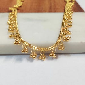 May include: Gold-toned anklet featuring a curb chain design with dangling clusters of small, round beads. The anklet is displayed on a white and light brown surface, showcasing its intricate details and reflective qualities.