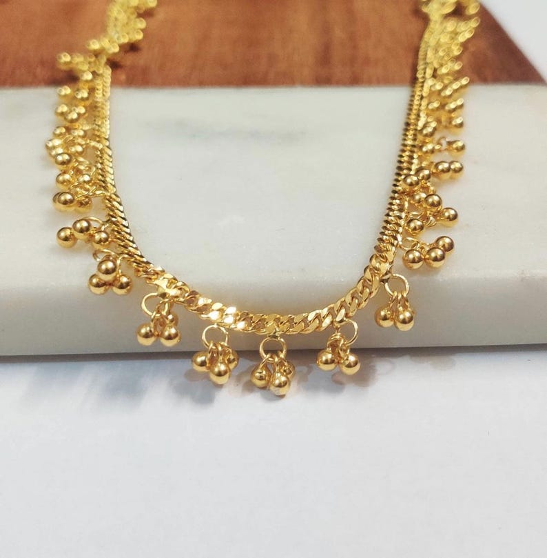 May include: Gold-toned anklet featuring a curb chain design with dangling clusters of small, round beads. The anklet is displayed on a white and light brown surface, showcasing its intricate details and reflective qualities.