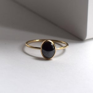 May include: A gold ring featuring a black oval gemstone. The ring has a thin, delicate band and a bezel setting that holds the polished black stone. The ring is set against a white background.