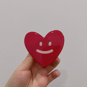 May include: A bright pink heart-shaped resin ornament with a white smiling face.