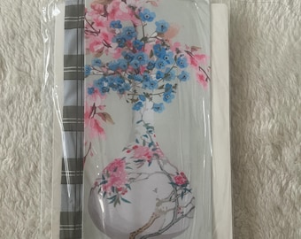 May include: A rectangular fabric panel featuring a light grey background with a central illustration of a white vase filled with blue and pink floral branches. The left side of the panel displays a vertical grey and white plaid pattern border.