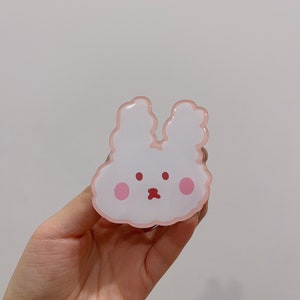 May include: A pink and white bunny-shaped phone grip with a cute cartoon face. The grip has a pink outline and two pink cheeks.