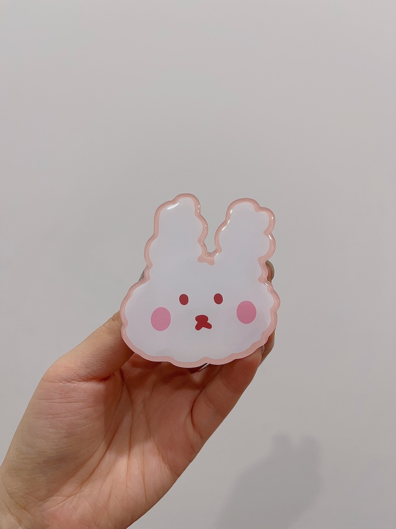 May include: A pink and white bunny-shaped phone grip with a cute cartoon face. The grip has a pink outline and two pink cheeks.