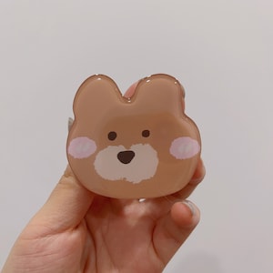 May include: A brown bear-shaped phone grip with a white face and pink cheeks. The grip is made of clear plastic and has a glossy finish.