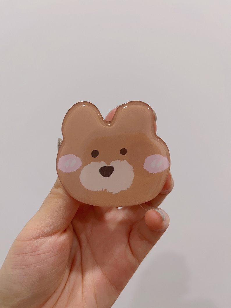May include: A brown bear-shaped phone grip with a white face and pink cheeks. The grip is made of clear plastic and has a glossy finish.