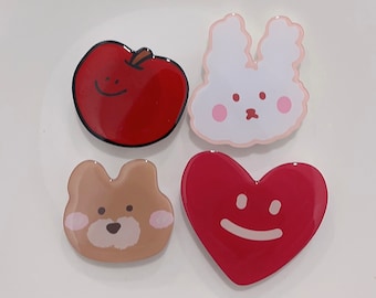 May include: Four colorful resin pins with cartoon faces. A red apple, a white bunny, a brown bear, and a red heart.