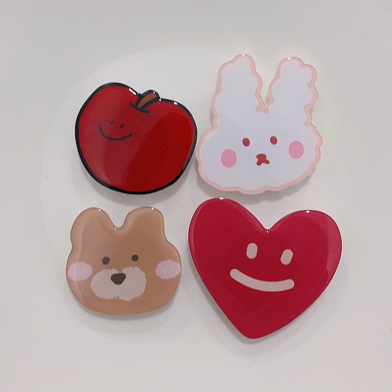 May include: Four colorful resin pins with cartoon faces. A red apple, a white bunny, a brown bear, and a red heart.