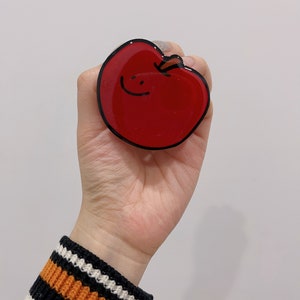 May include: A red apple phone grip with a black outline and a simple smiley face drawn on it.
