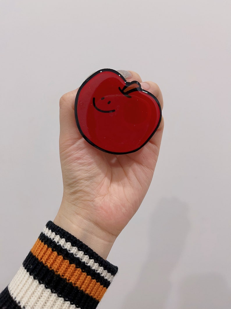 May include: A red apple phone grip with a black outline and a simple smiley face drawn on it.