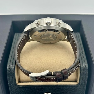 Puede incluir: Un reloj de pulsera de acero inoxidable con una correa de cuero marr&oacute;n. La esfera del reloj es visible, y la parte trasera del reloj tiene un dise&ntilde;o grabado. El reloj se muestra en una caja negra con un interior beige.
