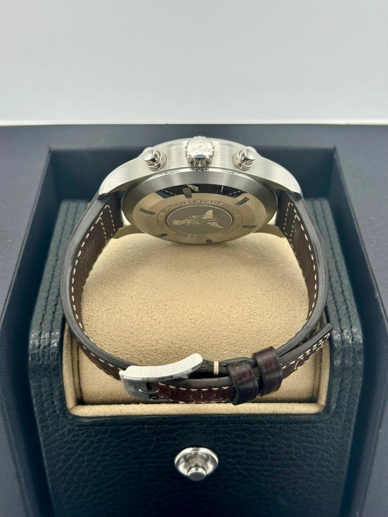 Puede incluir: Un reloj de pulsera de acero inoxidable con una correa de cuero marr&oacute;n. La esfera del reloj es visible, y la parte trasera del reloj tiene un dise&ntilde;o grabado. El reloj se muestra en una caja negra con un interior beige.