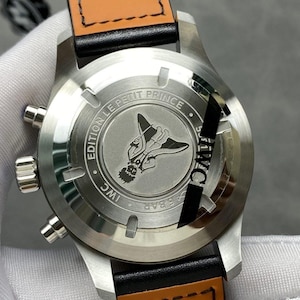 Puede incluir: Primer plano de un reloj de pulsera de acero inoxidable con una correa de cuero negro y una parte trasera de cuero marr&oacute;n. La parte trasera del reloj presenta una ilustraci&oacute;n grabada de una figura con alas y el texto "EDITION LE PETIT PRINCE" e "IWC".