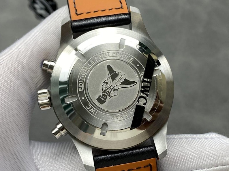 Puede incluir: Primer plano de un reloj de pulsera de acero inoxidable con una correa de cuero negro y una parte trasera de cuero marr&oacute;n. La parte trasera del reloj presenta una ilustraci&oacute;n grabada de una figura con alas y el texto "EDITION LE PETIT PRINCE" e "IWC".