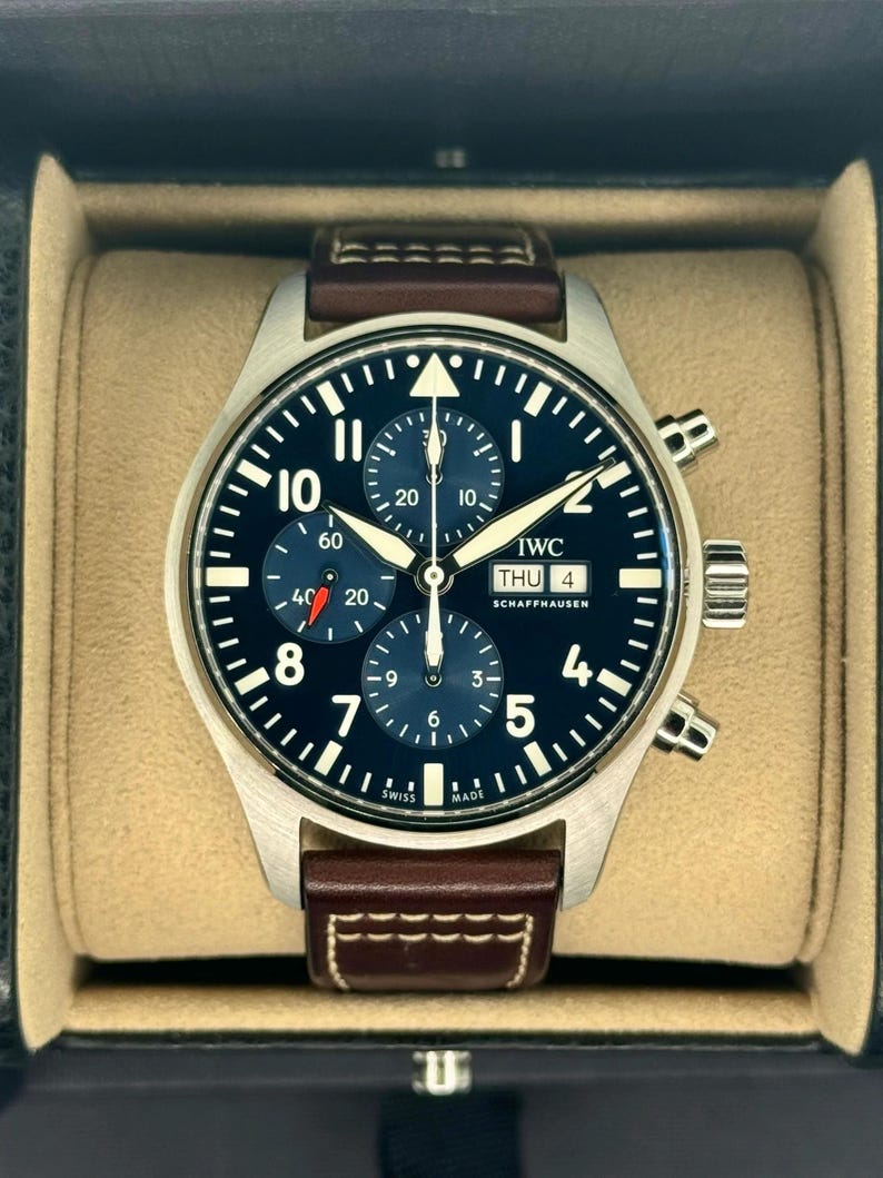 Puede incluir: Un reloj de pulsera de acero inoxidable con esfera azul oscuro, n&uacute;meros y agujas blancos. El reloj tiene una correa de cuero marr&oacute;n y se muestra en una caja beige. La esfera del reloj incluye el texto "IWC SCHAFFHAUSEN".