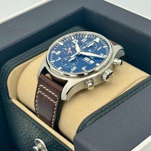 Puede incluir: Un reloj de pulsera de acero inoxidable con esfera azul y correa de cuero marr&oacute;n. El reloj presenta n&uacute;meros y agujas blancos, con la marca "IWC" visible en la esfera. El reloj se presenta en una caja de reloj negra y beige.