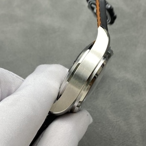 Puede incluir: Un reloj de pulsera plateado con una correa de cuero negro y marr&oacute;n. El reloj tiene un acabado de metal cepillado y un dise&ntilde;o elegante. El reloj est&aacute; sujeto por una mano enguantada.