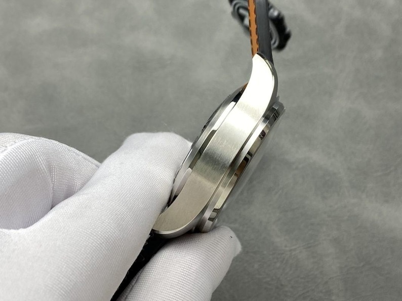 Puede incluir: Un reloj de pulsera plateado con una correa de cuero negro y marr&oacute;n. El reloj tiene un acabado de metal cepillado y un dise&ntilde;o elegante. El reloj est&aacute; sujeto por una mano enguantada.