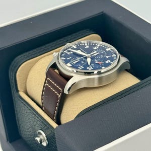 Puede incluir: Un reloj de pulsera de acero inoxidable con esfera azul oscuro y correa de cuero marr&oacute;n. El reloj se muestra en una caja negra y beige. La esfera del reloj tiene n&uacute;meros y agujas blancos.