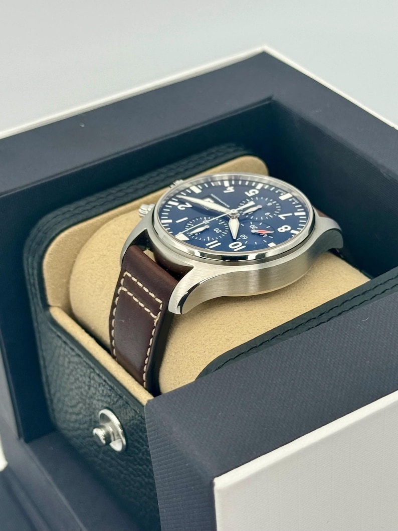 Puede incluir: Un reloj de pulsera de acero inoxidable con esfera azul oscuro y correa de cuero marr&oacute;n. El reloj se muestra en una caja negra y beige. La esfera del reloj tiene n&uacute;meros y agujas blancos.