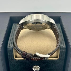 Puede incluir: Un reloj de pulsera de acero inoxidable con una correa de cuero marr&oacute;n. El reloj se muestra en una caja de reloj negra y beige. La correa tiene costuras blancas y una hebilla plateada. La esfera del reloj no es visible.