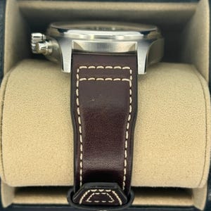 Puede incluir: Un reloj de pulsera de acero inoxidable con una correa de cuero marr&oacute;n. El reloj tiene una esfera redonda y una correa de cuero marr&oacute;n oscuro con costuras blancas. El reloj se muestra en una caja negra con interior beige.