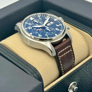 Puede incluir: Un reloj de pulsera de acero inoxidable con esfera azul y correa de cuero marr&oacute;n. El reloj presenta n&uacute;meros y agujas blancos, y se presenta en una caja de reloj negra y beige.