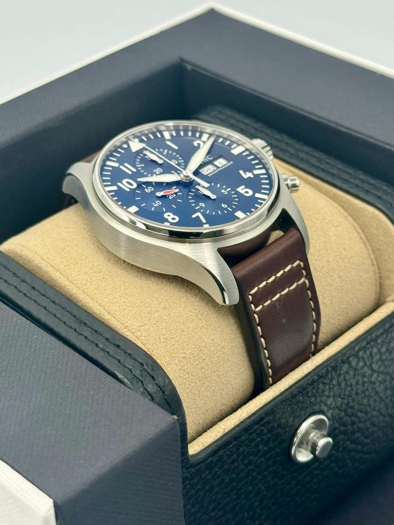 Puede incluir: Un reloj de pulsera de acero inoxidable con esfera azul y correa de cuero marr&oacute;n. El reloj presenta n&uacute;meros y agujas blancos, y se presenta en una caja de reloj negra y beige.