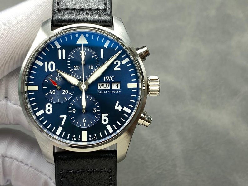 May include: A stainless steel wristwatch with a blue face and black leather band. The watch face features white numerals, sub-dials, and the text "IWC SCHAFFHAUSEN". The watch has a date display and two pushers on the side.