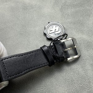 Puede incluir: Primer plano de una correa de reloj de cuero negro con hebilla plateada. La esfera del reloj es azul. Una etiqueta plateada con un logotipo est&aacute; sujeta a la correa.