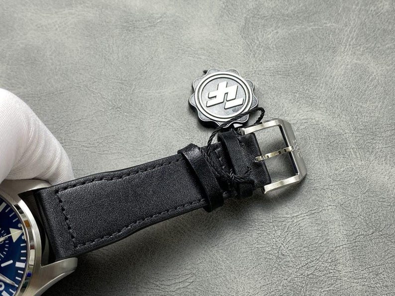 Puede incluir: Primer plano de una correa de reloj de cuero negro con hebilla plateada. La esfera del reloj es azul. Una etiqueta plateada con un logotipo est&aacute; sujeta a la correa.