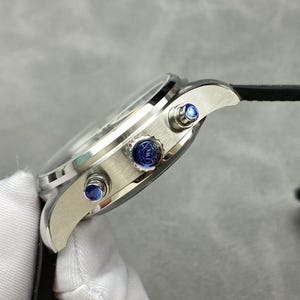 Puede incluir: Reloj de pulsera plateado con correa de cuero negro. El reloj tiene una esfera redonda con cristal transparente y detalles azules en los botones laterales. Vista lateral del reloj.