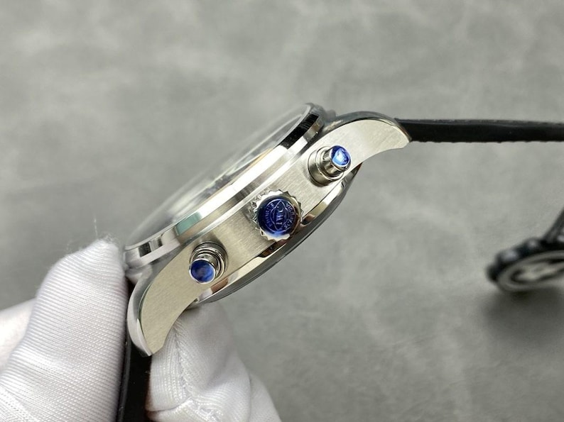 Puede incluir: Reloj de pulsera plateado con correa de cuero negro. El reloj tiene una esfera redonda con cristal transparente y detalles azules en los botones laterales. Vista lateral del reloj.