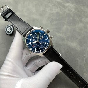 Puede incluir: Un reloj de pulsera de acero inoxidable con una correa de cuero negro. El reloj tiene una esfera azul con n&uacute;meros blancos y subesferas. El reloj est&aacute; siendo sostenido por una mano enguantada.