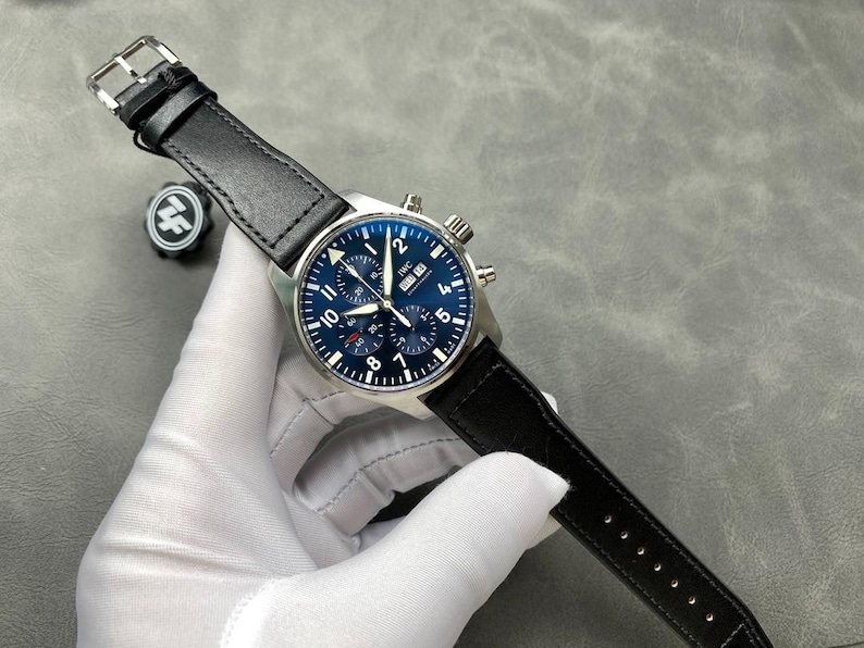Puede incluir: Un reloj de pulsera de acero inoxidable con una correa de cuero negro. El reloj tiene una esfera azul con n&uacute;meros blancos y subesferas. El reloj est&aacute; siendo sostenido por una mano enguantada.