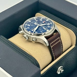 Puede incluir: Un reloj de pulsera de acero inoxidable con esfera azul y correa de cuero marr&oacute;n. El reloj se muestra en una caja negra y azul con interior beige. El reloj tiene funci&oacute;n cron&oacute;grafo y un peque&ntilde;o logotipo rojo.
