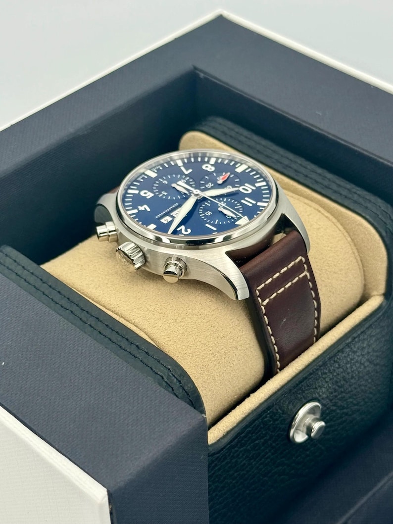 Puede incluir: Un reloj de pulsera de acero inoxidable con esfera azul y correa de cuero marr&oacute;n. El reloj se muestra en una caja negra y azul con interior beige. El reloj tiene funci&oacute;n cron&oacute;grafo y un peque&ntilde;o logotipo rojo.