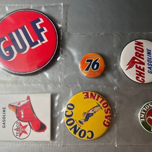 May include: A collection of six vintage gas station logo pins. The pins are round and feature various colors and designs, including red, white, blue, yellow, green, and orange. The logos include "Gulf", "Fire Chief", "Texaco", "Conoco", "Sinclair", "Union 76", and "Chevron".