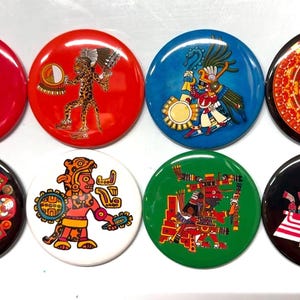 May include: A collection of eight round buttons featuring colorful Aztec-inspired designs. The buttons are in various colors, including red, blue, green, black, and white, with detailed illustrations of Aztec figures and symbols.