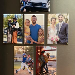 May include: A collection of rectangular magnets featuring various photographs. Images include a white SUV, a family portrait, a wedding photo, a baseball player, and couples in western attire. The magnets are arranged on a dark surface.