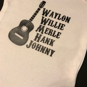 May include: White baby onesie with a black silhouette of a guitar and the names Waylon, Willie, Merle, Hank, and Johnny printed in black.