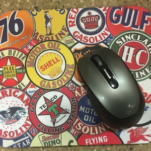 May include: A colorful mouse pad with a vintage design featuring various gas station logos and branding. The logos include 76, Shell, Sinclair, Gulf, Texaco, and Flying A. A gray computer mouse is on top of the mouse pad.