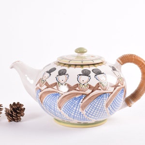 May include: A white ceramic teapot with a brown wicker handle. The teapot is decorated with a colorful pattern of people dancing in blue and brown dresses.