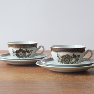May include: Two white teacups with brown floral designs and matching saucers. The cups and saucers have a brown band around the rim.