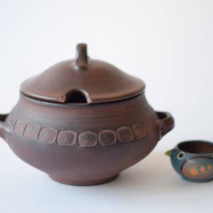 Può includere: Una zuppiera in ceramica marrone con coperchio e dettagli decorativi. La zuppiera ha un piccolo beccuccio sul coperchio. Un piccolo uccello in ceramica blu e marrone è accanto alla zuppiera.