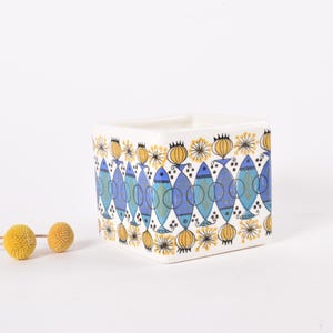 May include: Square ceramic container with a repeating pattern of blue fish and yellow floral designs on a white background. The container has a minimalist design and is suitable for various uses.