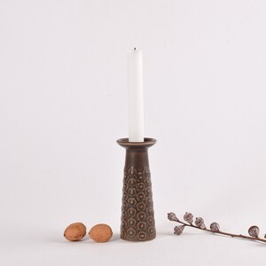 Puede incluir: Un candelero de cerámica marrón con un patrón floral y una vela blanca. El candelero está sobre una superficie blanca con dos nueces y una rama de eucalipto seco.