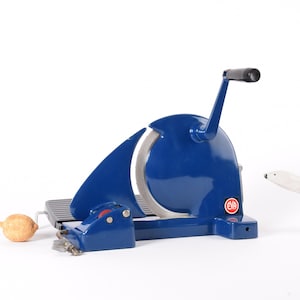 Puede incluir: Una cortadora azul vintage con un mango negro y una cuchilla plateada. También hay un pequeño limón y una figura de oso polar blanco. La cortadora tiene un logotipo rojo y blanco.